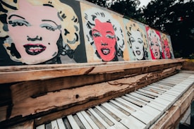 An artistic representation of famous pop art-style paintings featuring a woman's face on a wooden piano. The vibrant images are juxtaposed against the backdrop of an old, distressed piano with peeling paint and chipped keys. The setting appears outdoors with blurred greenery in the background.