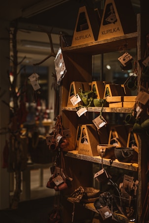 A wooden shelf in a store displays various handcrafted items such as cups, bowls, and utensils packaged in eco-friendly cardboard boxes. These items hang from hooks, showcasing earthy tones and rustic designs, with natural lighting emphasizing the warm atmosphere.