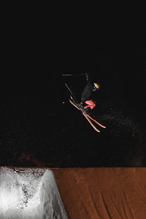 A skier is airborne, performing a trick under dark night conditions. The bright ski jump contrasts sharply with the dark background.