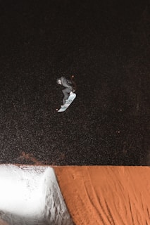 A snowboarder in mid-air performing a trick against a dark, snowy backdrop. The snowboarder is wearing a helmet and dark clothing, with the snowboard angled beneath them. The image captures the dynamic movement of the trick being executed over a snow ramp with visible tracks.