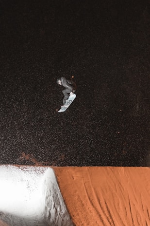 A snowboarder in mid-air performing a trick against a dark, snowy backdrop. The snowboarder is wearing a helmet and dark clothing, with the snowboard angled beneath them. The image captures the dynamic movement of the trick being executed over a snow ramp with visible tracks.
