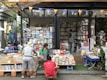 A street bookstore with shelves stacked high with a variety of books. People are browsing through the selections, with one person wearing a hat and another in a green jacket. The books are organized both inside the shop and outside, on tables and stands. A string of triangular flags adds a festive touch to the scene.
