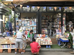 A street bookstore with shelves stacked high with a variety of books. People are browsing through the selections, with one person wearing a hat and another in a green jacket. The books are organized both inside the shop and outside, on tables and stands. A string of triangular flags adds a festive touch to the scene.