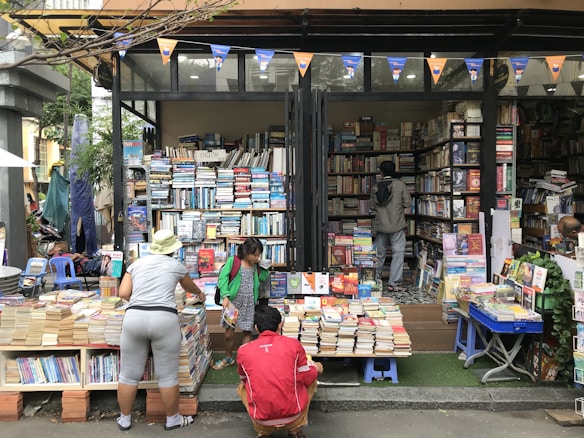 A street bookstore with shelves stacked high with a variety of books. People are browsing through the selections, with one person wearing a hat and another in a green jacket. The books are organized both inside the shop and outside, on tables and stands. A string of triangular flags adds a festive touch to the scene.