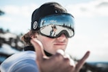 A person wearing ski goggles and a black cap makes a hand gesture commonly associated with surfing or good vibes. The reflective surface of the goggles shows a snowy landscape with trees and a clear blue sky.