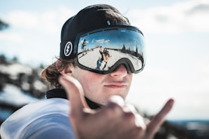 A person wearing ski goggles and a black cap makes a hand gesture commonly associated with surfing or good vibes. The reflective surface of the goggles shows a snowy landscape with trees and a clear blue sky.