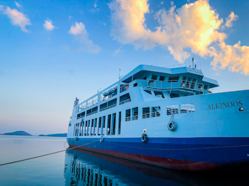 A passenger ferry named 'ALKINOOS' is docked, resting on calm waters. The vessel has a white and blue color scheme, with visible life rings and a series of large windows along its side. The sky above is mostly clear with a few fluffy clouds illuminated by the golden hues of sunset.