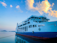A passenger ferry named 'ALKINOOS' is docked, resting on calm waters. The vessel has a white and blue color scheme, with visible life rings and a series of large windows along its side. The sky above is mostly clear with a few fluffy clouds illuminated by the golden hues of sunset.