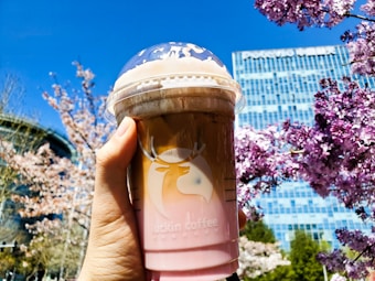 A hand holding a branded cup of iced coffee with a plastic lid, set against a background of blooming cherry blossoms and a clear blue sky. A modern building is visible in the background.