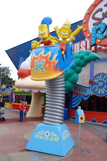 A whimsical theme park ride featuring characters resembling a cartoon family sitting in a vibrant car atop a spring-like pedestal. The ride is surrounded by colorful signage and decorative elements, with people in the background.