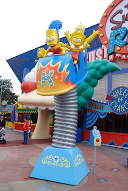 A whimsical theme park ride featuring characters resembling a cartoon family sitting in a vibrant car atop a spring-like pedestal. The ride is surrounded by colorful signage and decorative elements, with people in the background.