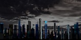 A futuristic cityscape featuring a skyline with a variety of tall, modern skyscrapers under a dramatic, cloud-filled sky. The buildings are illuminated with lights and digital screens, some displaying logos or advertisements. The setting appears to be at dusk or night, giving a vibrant yet somewhat eerie atmosphere.