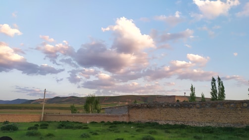 A scenic landscape featuring a brick wall in the foreground, lush green grass, and a few tall trees. Beyond the wall, rolling hills stretch across the horizon under a sky filled with fluffy white clouds tinged with soft pink and orange hues from the setting sun.