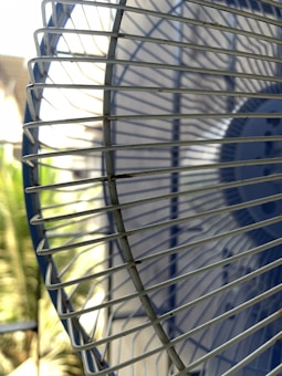 A close-up view of a metallic fan grill with a blurred background. The image highlights the intersecting wires and shadows cast by the grill, creating a pattern of lines and curves. Sunlight is visible, giving the metal a reflective sheen.