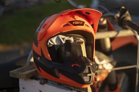 A bright orange and black motocross helmet is placed on a wooden surface, partially covering the handlebars and seat of a motorcycle in the background. The design features angular shapes and prominent vents, with a brand logo visible on the top. The lighting is warm, suggesting sunset or sunrise.
