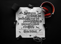 A black and white typography print on a white paper features ornate, Gothic-style lettering. Surrounding the paper are items including a pair of red headphones, a black Bluetooth speaker, a toy car, and a rope. Small bat illustrations can be seen on the paper, enhancing the dark theme.