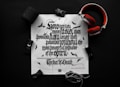 A black and white typography print on a white paper features ornate, Gothic-style lettering. Surrounding the paper are items including a pair of red headphones, a black Bluetooth speaker, a toy car, and a rope. Small bat illustrations can be seen on the paper, enhancing the dark theme.