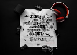 A black and white typography print on a white paper features ornate, Gothic-style lettering. Surrounding the paper are items including a pair of red headphones, a black Bluetooth speaker, a toy car, and a rope. Small bat illustrations can be seen on the paper, enhancing the dark theme.