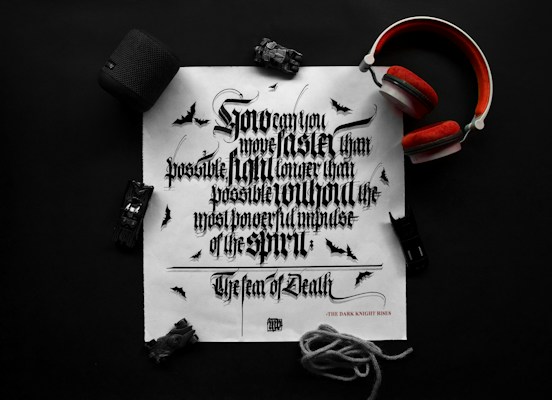 A black and white typography print on a white paper features ornate, Gothic-style lettering. Surrounding the paper are items including a pair of red headphones, a black Bluetooth speaker, a toy car, and a rope. Small bat illustrations can be seen on the paper, enhancing the dark theme.