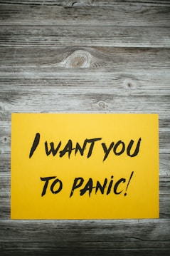 A yellow paper with bold black text stating 'I want you to panic!' is placed on a rustic wooden surface with a grayish-brown weathered texture.