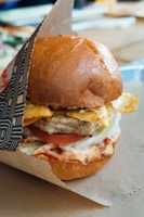 A close-up of a wrapped burger with a browned bun, crispy chips, a juicy chicken patty, sliced tomato, lettuce, cheese, and creamy sauces layers inside.