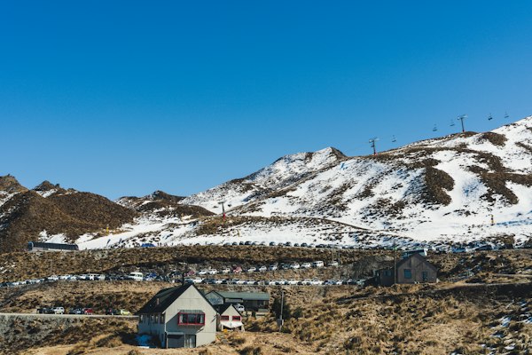 Coronet Peak base building and car park with chairlift towers climbing a snowy tussock ridge under a blue sky