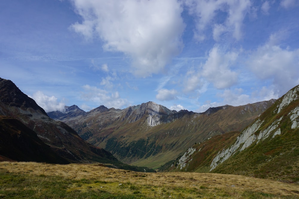 green grass field near brown mountains under white clouds and blue sky during daytime