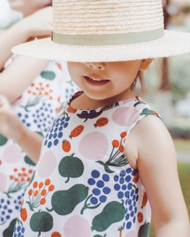 A child is wearing a dress with colorful, abstract fruit and vegetable patterns. The child is partially obscured by a straw hat with a green ribbon that covers their eyes. The background is blurred, suggesting an outdoor setting.