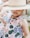 A child is wearing a dress with colorful, abstract fruit and vegetable patterns. The child is partially obscured by a straw hat with a green ribbon that covers their eyes. The background is blurred, suggesting an outdoor setting.