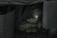A person wearing a green uniform with a patch on the arm, holding a hat, is seated behind large protective shields. The setting is dimly lit and appears to be inside a vehicle or tent, with a military or tactical theme.