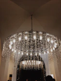 An elaborate chandelier with multiple lit bulbs is adorned with cascading strings of fairy lights, creating a warm and inviting ambiance. The ceiling is arched, and there are hints of additional lighting visible in the background through an archway.