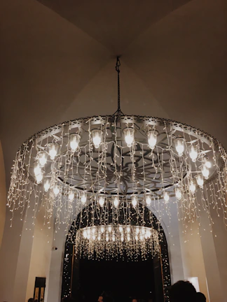 An elaborate chandelier with multiple lit bulbs is adorned with cascading strings of fairy lights, creating a warm and inviting ambiance. The ceiling is arched, and there are hints of additional lighting visible in the background through an archway.