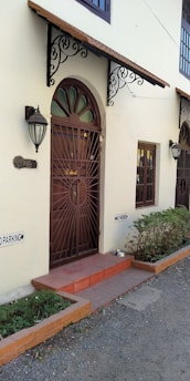 A residential entrance featuring a decorative metal door under an ornate awning. Two outdoor wall lamps flank the entrance. There are 'No Parking' signs on the wall and a small potted plant next to the door. The pathway is outlined with low brick borders and the exterior is painted in a light cream color.