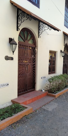 A residential entrance featuring a decorative metal door under an ornate awning. Two outdoor wall lamps flank the entrance. There are 'No Parking' signs on the wall and a small potted plant next to the door. The pathway is outlined with low brick borders and the exterior is painted in a light cream color.