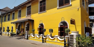 A vibrant yellow colonial-style building with large windows and decorative ironwork. The structure features elegant archways and is bordered by a low white fence with carefully trimmed shrubs. A street in front is lined with cobblestones, and a person is standing near the entrance, adding a touch of life to the scene. The blue sky and overhead power lines complete the urban setting.