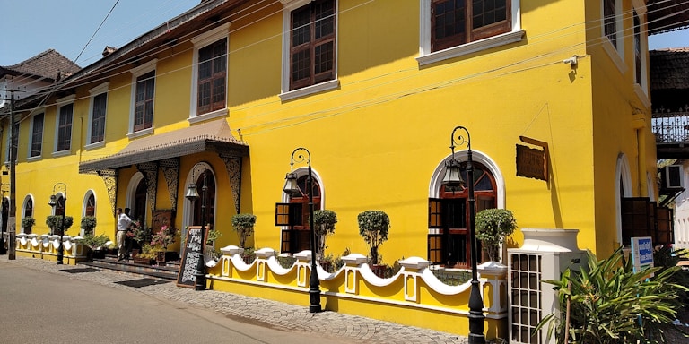 A vibrant yellow colonial-style building with large windows and decorative ironwork. The structure features elegant archways and is bordered by a low white fence with carefully trimmed shrubs. A street in front is lined with cobblestones, and a person is standing near the entrance, adding a touch of life to the scene. The blue sky and overhead power lines complete the urban setting.