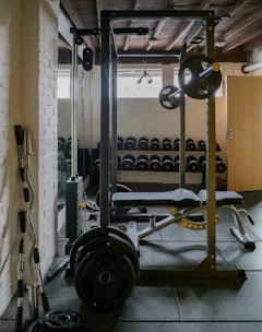 A well-equipped gym with a focus on weightlifting. There is a squat rack with various weighted plates attached. In the background, rows of dumbbells are neatly organized on racks, and a bench press setup is visible. The room features white brick walls and a wooden ceiling, contributing to an industrial vibe.