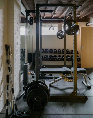 A well-equipped gym with a focus on weightlifting. There is a squat rack with various weighted plates attached. In the background, rows of dumbbells are neatly organized on racks, and a bench press setup is visible. The room features white brick walls and a wooden ceiling, contributing to an industrial vibe.