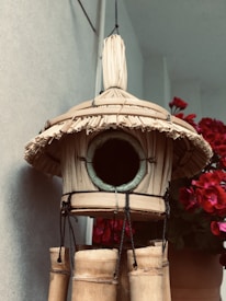 A handcrafted birdhouse made of straw and bamboo, featuring a circular entrance. It is suspended with black ropes. In the background, there are vibrant red flowers in a pot.