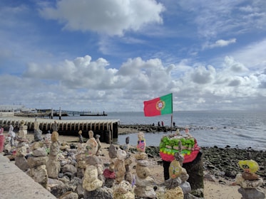 A seaside scene with a rocky shore decorated with painted rocks and sculptures, featuring a prominently displayed Portuguese flag waving in the breeze. The sky is partly cloudy with patches of blue, and a pier extends into the water.