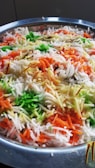 A close-up view of a dish featuring long-grain rice mixed with vibrant colors. Green, orange, and white colors are prominent, suggesting the use of spices or food coloring. The rice appears fluffy and is served in a metal container.