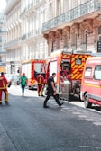 Several fire trucks and emergency personnel in uniforms are present on a city street lined with ornate buildings. A few pedestrians can also be seen walking nearby, and metal barricades are set up, suggesting a controlled or restricted area.