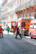 Several fire trucks and emergency personnel in uniforms are present on a city street lined with ornate buildings. A few pedestrians can also be seen walking nearby, and metal barricades are set up, suggesting a controlled or restricted area.
