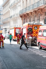 Several fire trucks and emergency personnel in uniforms are present on a city street lined with ornate buildings. A few pedestrians can also be seen walking nearby, and metal barricades are set up, suggesting a controlled or restricted area.