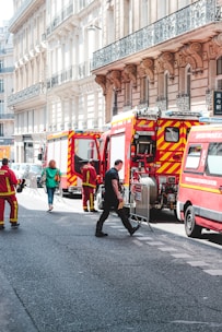 Several fire trucks and emergency personnel in uniforms are present on a city street lined with ornate buildings. A few pedestrians can also be seen walking nearby, and metal barricades are set up, suggesting a controlled or restricted area.