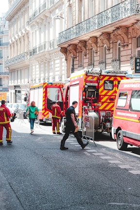 Several fire trucks and emergency personnel in uniforms are present on a city street lined with ornate buildings. A few pedestrians can also be seen walking nearby, and metal barricades are set up, suggesting a controlled or restricted area.