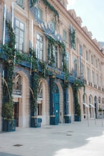 An elegant building facade covered with lush green vines and blue decorative elements. The architecture features large arched windows and detailed stonework, conveying a luxurious and classic style. The entrance prominently displays signage with ornate lettering, and the building is situated on a spacious, sunlit street.
