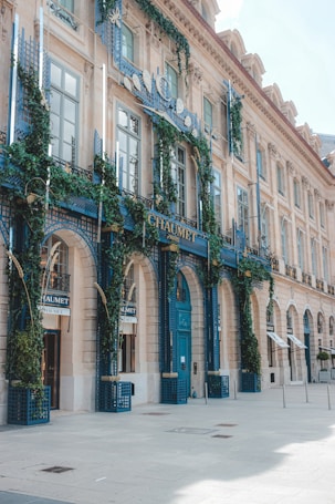 An elegant building facade covered with lush green vines and blue decorative elements. The architecture features large arched windows and detailed stonework, conveying a luxurious and classic style. The entrance prominently displays signage with ornate lettering, and the building is situated on a spacious, sunlit street.