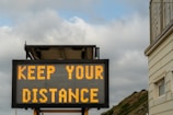 A digital road sign displaying the message 'KEEP YOUR DISTANCE' against a backdrop of cloudy skies and a building edge. The sign is illuminated with bright yellow text, suggesting a cautionary message to onlookers.