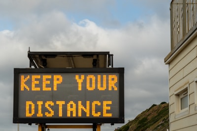 A digital road sign displaying the message 'KEEP YOUR DISTANCE' against a backdrop of cloudy skies and a building edge. The sign is illuminated with bright yellow text, suggesting a cautionary message to onlookers.